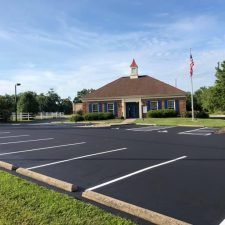 Freshly sealcoated commercial parking lot with a smooth, black surface, featuring bright, well-defined parking lines and surrounded by a row of neatly landscaped greenery and a modern office building in the background